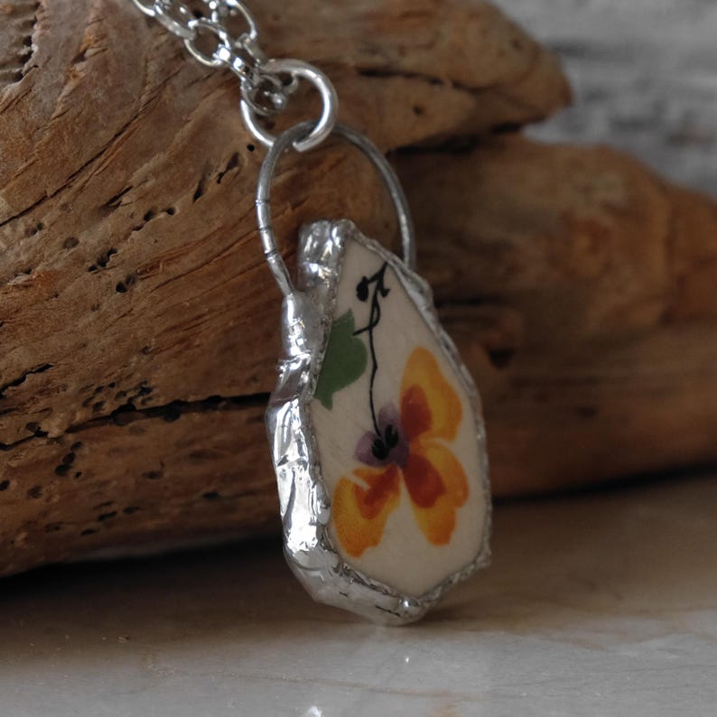 Puede incluir: Collar plateado con colgante floral. El colgante tiene un fondo blanco con una flor amarilla y naranja, una hoja verde y detalles negros. El colgante est&aacute; enmarcado en un marco de metal plateado. Joyer&iacute;a.
