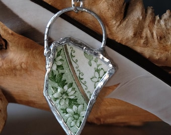 Puede incluir: Collar con colgante artesanal que presenta un fragmento de porcelana vintage con un diseño floral verde. La porcelana está engarzada en un marco de metal plateado y cuelga de una fina cadena. Una pieza única con encanto vintage.
