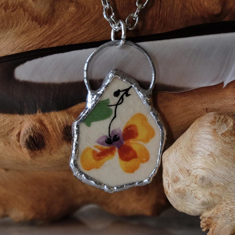 Puede incluir: Un collar con colgante hecho a mano con un dise&ntilde;o floral. El colgante tiene una forma &uacute;nica, con un marco de metal plateado. El dise&ntilde;o floral tiene tonos amarillos, naranjas y morados. El collar est&aacute; en una cadena plateada.