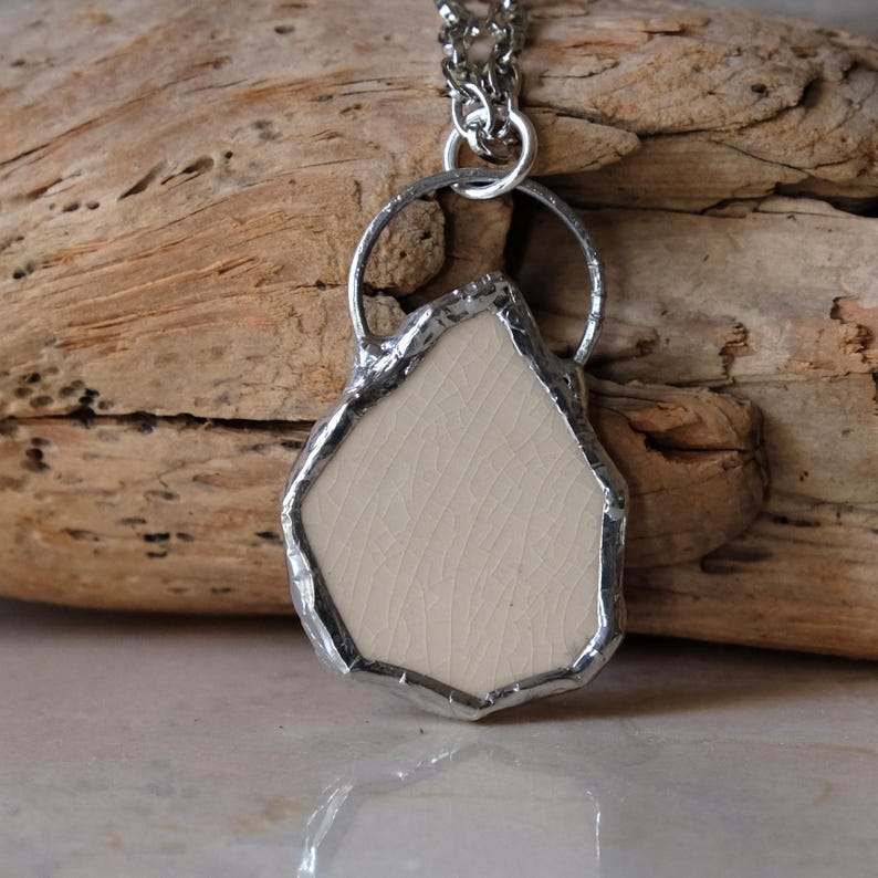 Puede incluir: Un collar con colgante artesanal con una pieza de cer&aacute;mica de color crema, de forma irregular, enmarcada en un marco de metal plateado. El colgante cuelga de una delicada cadena plateada, mostrando un dise&ntilde;o art&iacute;stico &uacute;nico.
