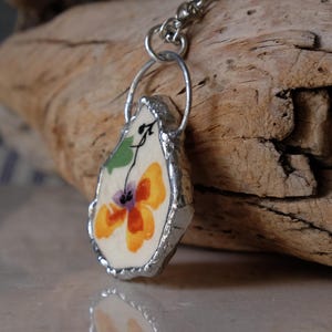 Puede incluir: Un colgante hecho a mano con un dise&ntilde;o floral. El colgante tiene un marco de metal plateado y una flor pintada con tonos naranja, morado y verde. Cuelga de una cadena plateada, mostrando una pieza art&iacute;stica &uacute;nica.