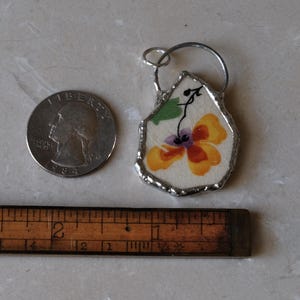 Puede incluir: Un colgante hecho a mano con un dise&ntilde;o floral en tonos amarillos, naranjas y morados. El colgante est&aacute; enmarcado con metal plateado e incluye un peque&ntilde;o lazo para colgarlo. Una moneda y una regla est&aacute;n junto al colgante.