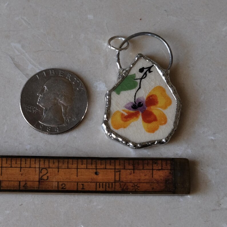 Puede incluir: Un colgante hecho a mano con un dise&ntilde;o floral en tonos amarillos, naranjas y morados. El colgante est&aacute; enmarcado con metal plateado e incluye un peque&ntilde;o lazo para colgarlo. Una moneda y una regla est&aacute;n junto al colgante.