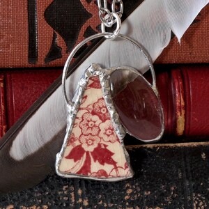 Pode incluir: Um colar com pingente de prata feito à mão, com um pedaço triangular de cerâmica com estampa floral vermelha e branca e uma pedra ovalada de cor bordô. O pingente pende de uma delicada corrente de prata, mostrando um design artístico único.