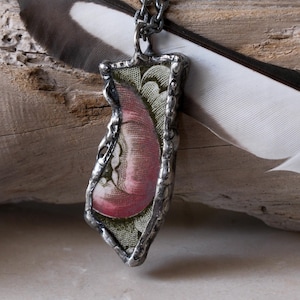 Pode incluir: Um colar com pingente artesanal com um design único. O pingente apresenta uma inserção estampada em rosa e verde, emoldurada em uma estrutura de metal prateado. O colar é exibido com uma pena e um pedaço de madeira.