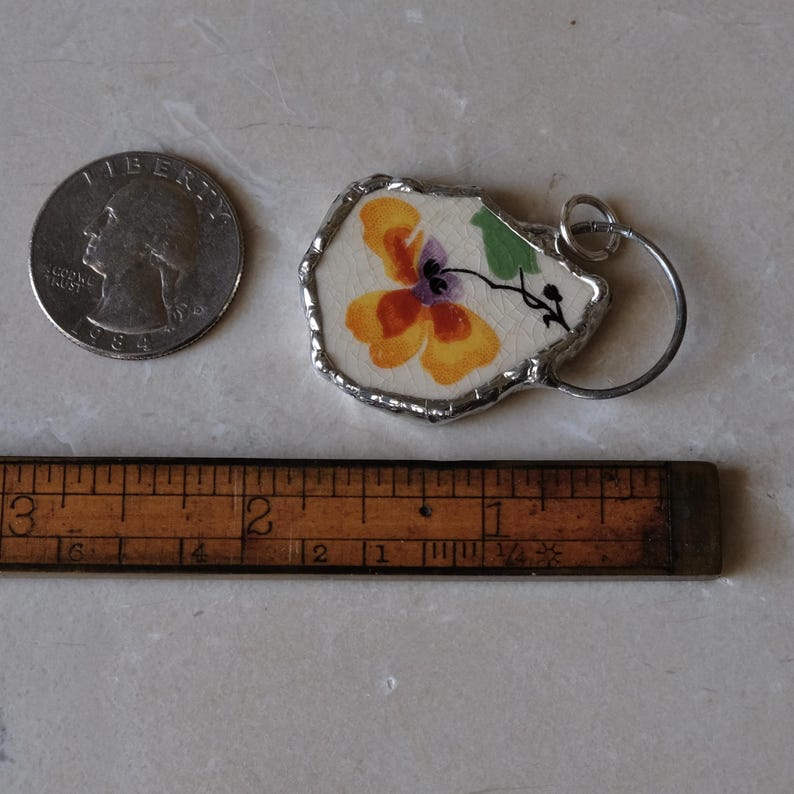 Puede incluir: Un colgante hecho a mano con un dise&ntilde;o floral pintado en tonos naranja, amarillo y morado. El colgante est&aacute; enmarcado con metal plateado e incluye un peque&ntilde;o anillo de metal. Un cuarto de d&oacute;lar y una regla de madera est&aacute;n al lado.
