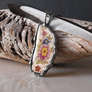 May include: A handcrafted pendant necklace featuring a floral design. The pendant is made from a piece of vintage china with a colorful floral pattern, encased in a silver-colored metal frame. The necklace is displayed on a piece of driftwood with a white feather.