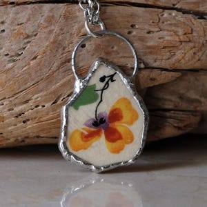 Puede incluir: Un collar con colgante hecho a mano con un colgante de porcelana en forma de l&aacute;grima. El colgante tiene un dise&ntilde;o floral en naranja, morado y verde, enmarcado en metal plateado y cuelga de una delicada cadena.