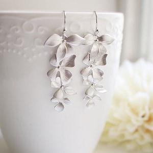 May include: Silver-toned floral drop earrings. Each earring features a cascade of delicate, flower-shaped elements. The earrings are displayed against a white background, with a white ceramic cup and a soft, white flower in the background.