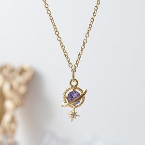 Puede incluir: Un collar de oro con un colgante de piedra preciosa púrpura que se asemeja a un planeta con un anillo a su alrededor y una pequeña estrella debajo.