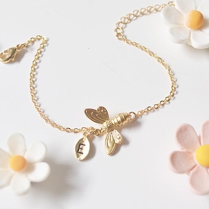 Puede incluir: Una pulsera dorada con un dije de abeja y un colgante inicial, rodeada de flores de margarita decorativas en blanco y rosa. La pulsera es una joya delicada.