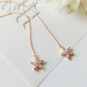 Puede incluir: Un par de pendientes de cadena dorada con un pequeño dije en forma de flor al final de cada cadena. Los dijes están hechos de diamantes de imitación transparentes.