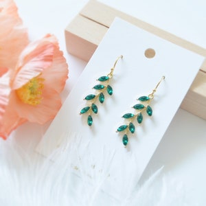 May include: A pair of gold earrings with emerald green crystal leaves. The earrings are dangle style and have a delicate, feminine look.