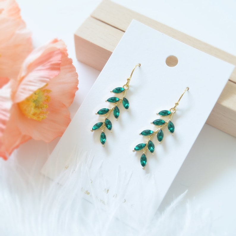 May include: A pair of gold earrings with emerald green crystal leaves. The earrings are dangle style and have a delicate, feminine look.