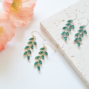 May include: A pair of dangle earrings with a leafy design. The earrings are made of gold and feature emerald green crystals.