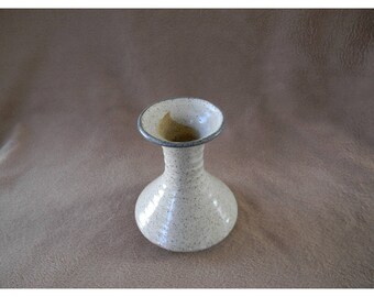 May include: A small, white ceramic vase with a speckled glaze and a dark gray rim. The vase has a rounded base and a narrow neck.