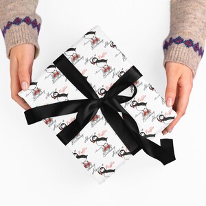 Puede incluir: Un regalo envuelto con un lazo de satén negro. El papel de regalo es blanco con un patrón repetido de frailecillos, flores y las palabras "Happy Birthday Puffin" en escritura roja y negra. El regalo es sostenido por una persona que lleva un suéter beige.