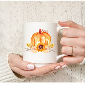 May include: White ceramic mug with a watercolor design of a pumpkin, sunflower, and leaves. The pumpkin is orange and the sunflower is yellow with brown center. The leaves are brown and green.