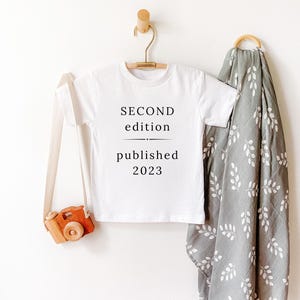 May include: White baby onesie with black text that reads "SECOND edition published 2023". The onesie is hanging on a wooden hanger. A wooden toy camera and a gray muslin swaddle blanket with white leaf pattern are in the background.