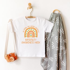 Op de afbeelding: Wit T-shirt met een regenboogontwerp en de tekst "In April We Wear Orange" en "Infertility Awareness Week". De regenboog is gemaakt van ananas.