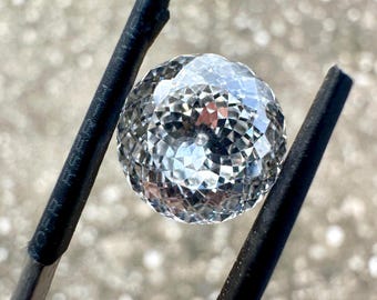Può includere: Una gemma rotonda, trasparente e sfaccettata, tenuta da una pinzetta in metallo nero. La pietra presenta un motivo complesso e simmetrico di sfaccettature triangolari e a diamante che riflettono la luce, creando un aspetto brillante.