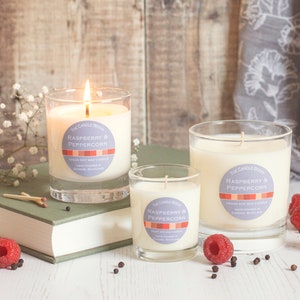 May include: Three glass candle jars with a white wax base and a purple and red label that reads "Raspberry & Peppercorn" on a white wooden surface with fresh raspberries and black peppercorns.