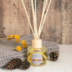 May include: A glass diffuser bottle with wooden reeds, labelled "Warming Honey & Tobacco." The bottle sits on a grey knitted blanket, with yellow flowers and pine cones. The background is a wooden surface.