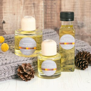 May include: Three glass bottles of diffuser oil with wooden stoppers. The bottles are filled with a yellow liquid and have labels that read "Warming Honey & Tobacco Diffuser".