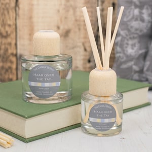 May include: Two glass diffuser bottles with wooden caps and reeds. The bottles are labelled "The Candle Bothe" and "Haar Over The Tay" diffuser. The bottles are sitting on a green book.