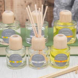 May include: Six glass diffuser bottles with wooden caps and reeds. The bottles are filled with clear liquid and have labels with different scents, including "Sunny Dundee", "Dundee Marmalade", and "Haar Over The Tay".