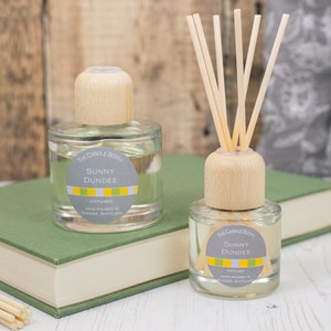 May include: Two glass diffuser bottles with wooden caps and reeds. The bottles are filled with a clear liquid and have labels that say "The Candle Botan", "Sunny Dundee", and "Diffuser".