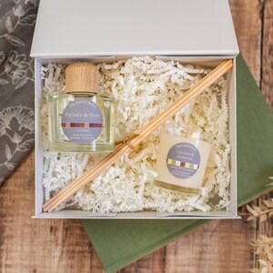 May include: A white gift box filled with white shredded paper, containing a clear glass diffuser bottle with a wooden lid and a small glass candle. The diffuser bottle has a label that reads "Peony & Oud", and the candle has a label that reads "Cedar & Jasmine".
