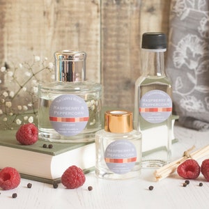 May include: Three glass bottles of diffuser oil with a raspberry and peppercorn scent. The bottles are clear and have a silver, gold, and black lid. The bottles are sitting on a white surface with a book, raspberries, and peppercorns.
