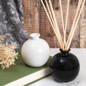May include: Two round ceramic vases, one white and one black, with a book and dried plant in the background. The black vase has wooden sticks inserted into it.