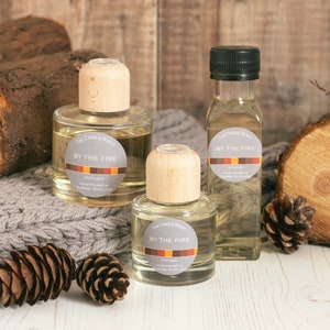 May include: Three glass diffuser bottles and a small bottle of oil are arranged on a wooden surface with a grey knitted blanket, pine cones, and wood logs. The bottles have wooden tops and labels that read "BY THE FIRE".