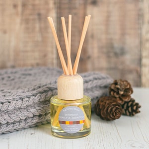May include: A clear glass bottle diffuser with a wooden lid and natural reeds. The bottle contains a yellow liquid and has a label that reads "Warming Honey & Tobacco" with a decorative stripe pattern. The diffuser is sitting on a white surface with a grey knitted blanket and pine cones.