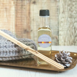 May include: A clear glass bottle of diffuser oil with a black cap and a label that reads "The Candle Room Warming Honey & Tobacco". The bottle is sitting on a gold tray with wooden diffuser sticks and a pine cone.