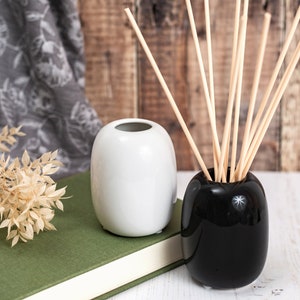 May include: Two ceramic vases, one white and one black, with a set of wooden diffuser sticks in the black vase. The vases are on a white surface with a green book and a dried plant in the foreground.