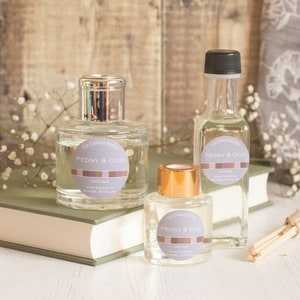 May include: Three glass bottles of diffuser oil with a gold and silver lid. The bottles are filled with a clear liquid and have a label that says "Peony & Oud" in a white circle with a pink and purple stripe. The bottles are sitting on a green book with a white surface underneath.