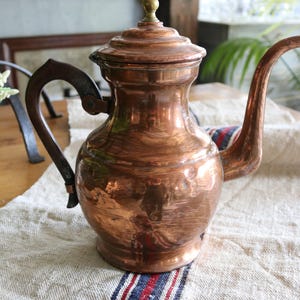 May include: An antique copper teapot with a curved spout and a dark handle. The teapot has a lid with a small brass knob. It rests on a linen cloth with red, white, and blue stripes. The teapot is a warm, antique colour.