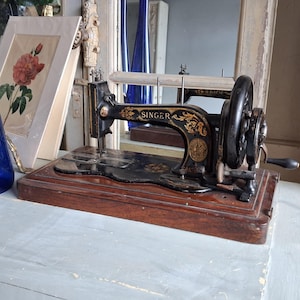 May include: A vintage black Singer sewing machine featuring intricate gold floral decals and a hand crank mechanism. The machine is mounted on a rectangular wooden base and displays the Singer brand name clearly on the side of the metal frame.
