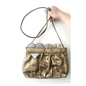 Könnte beinhalten: Eine goldfarbene Handtasche mit herzförmigem Design und silberfarbenem Besatz. Die Tasche hat einen langen Riemen und ist aus einem strukturierten Stoff gefertigt.