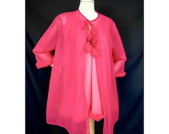 May include: A sheer, hot pink vintage robe with a matching slip. The robe features a tie closure with three decorative pom-poms and ruffled trim on the sleeves and hem. The garment is displayed on a mannequin against a black backdrop.