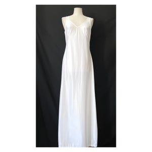 Op de afbeelding: Een witte, mouwloze, lange slipdress met een V-hals. De jurk is gemaakt van een gladde, glanzende stof en hangt recht van de schouders tot de vloer. De jurk wordt gepresenteerd op een mannequin tegen een zwarte achtergrond.