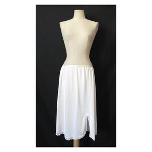 May include: A white slip skirt displayed on a mannequin. The skirt is knee-length with a slit on the side and delicate lace trim along the hem and slit. The fabric appears smooth and lightweight, suitable for layering.