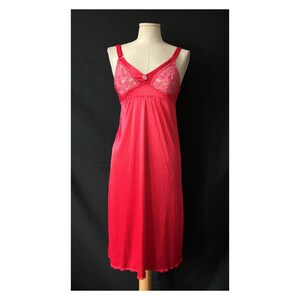 May include: A vintage red slip dress with a V-neck and lace detailing on the bodice. The dress has adjustable straps and a flowing, knee-length skirt. The slip is displayed on a mannequin against a black background.