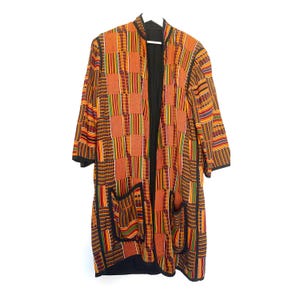May include: A long, open-front jacket with a patchwork design in shades of orange, brown, and black. The jacket features a black lining, two front pockets, and a collar. The sleeves are three-quarter length, and the jacket is hanging on a white hanger.