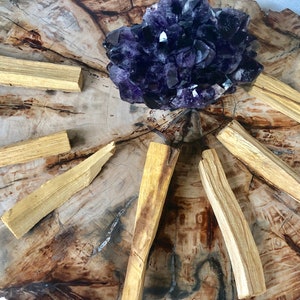 May include: A collection of Palo Santo sticks arranged in a circle around a large amethyst crystal on a wooden surface.