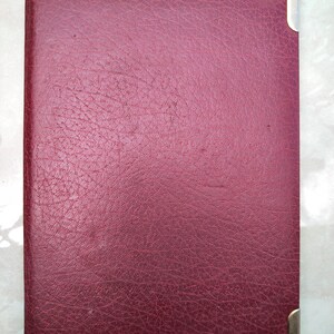 May include: A rectangular book with a deep burgundy, textured cover. The front cover features polished gold-tone metal corner protectors on the top right and bottom right corners, providing a contrast to the dark red, grained surface of the book.