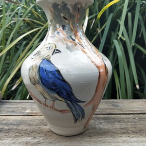 May include: A cream-coloured ceramic vase featuring a hand-painted design of a bird with a yellow head and blue wings perched on a branch. The artwork includes a brown tree trunk and muted green foliage accents, with a wide rim and tapered base.